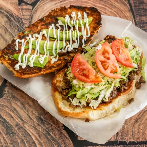 An open El Güero Canelo torta with meat, lettuce, tomatoes, avocado, and crema on toasted bread.