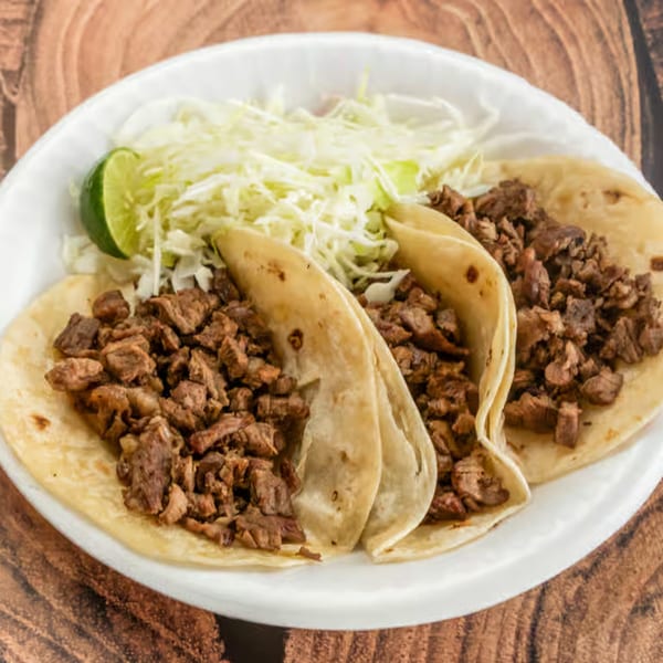 Three carne asada tacos with shredded cabbage and a lime wedge on a white plate.