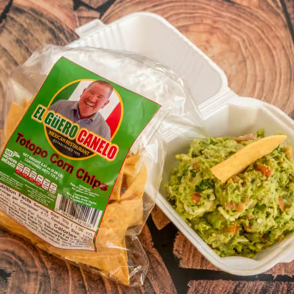 El Güero Canelo Totopo Corn Chips bag next to a white container of guacamole with a chip.