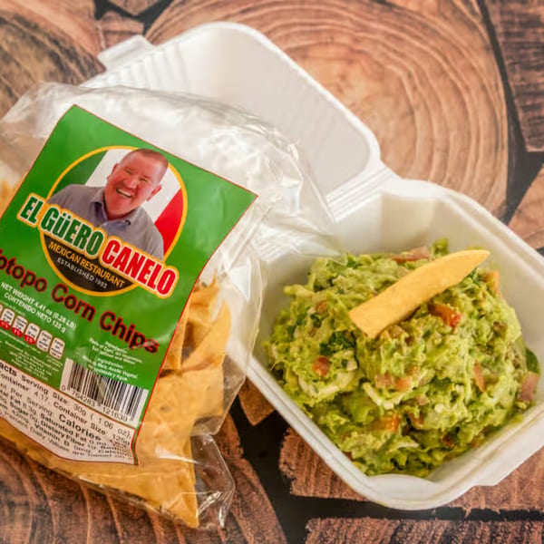 El Güero Canelo corn chips next to a container of guacamole with a chip on top.