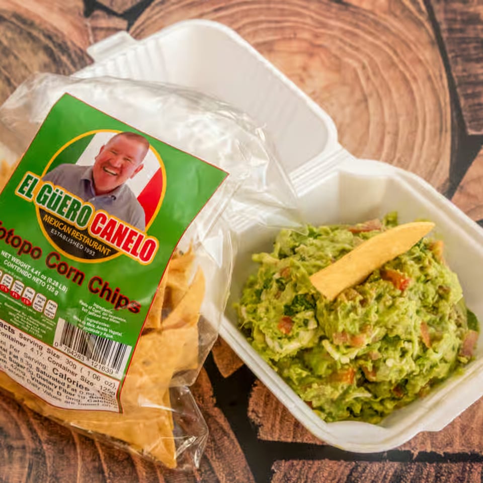 El Güero Canelo corn chips next to a container of guacamole with a chip on top.